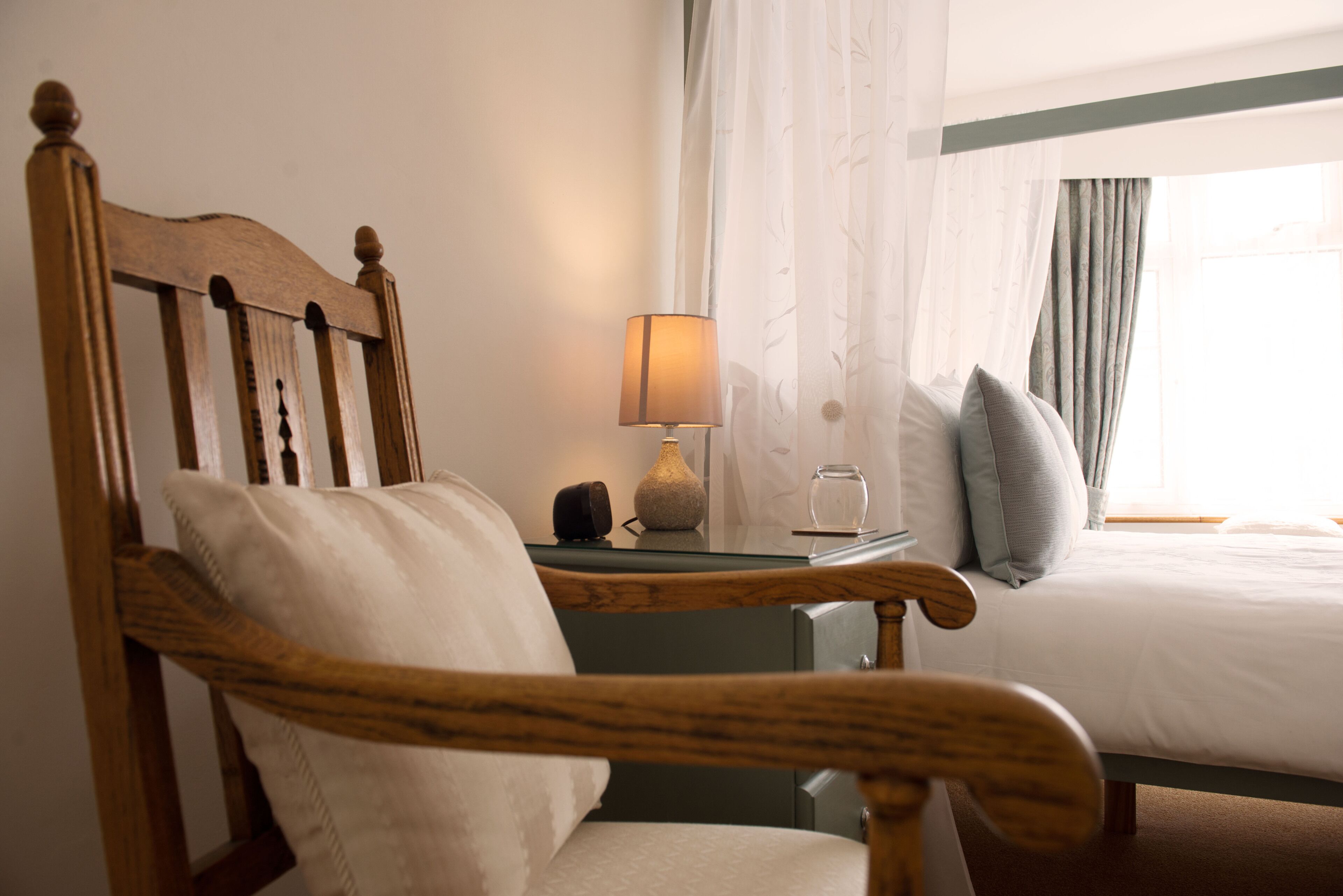 A wooden chair with a cushion, a bedside table with a lamp, and a bed with pillows.
