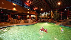 Une piscine intérieure avec une personne sur un flotteur, de l’eau claire et une vue sur le ciel à travers de grandes fenêtres.