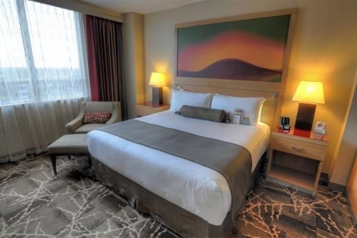 Hotelkamer met een groot bed, twee bedlampen, een stoel, een schilderij aan de muur en een raam met gordijnen.