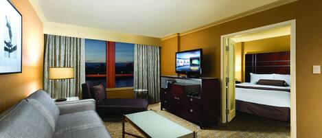 A hotel room with a sofa, a TV, a desk, a bed, and a view of the city at night.