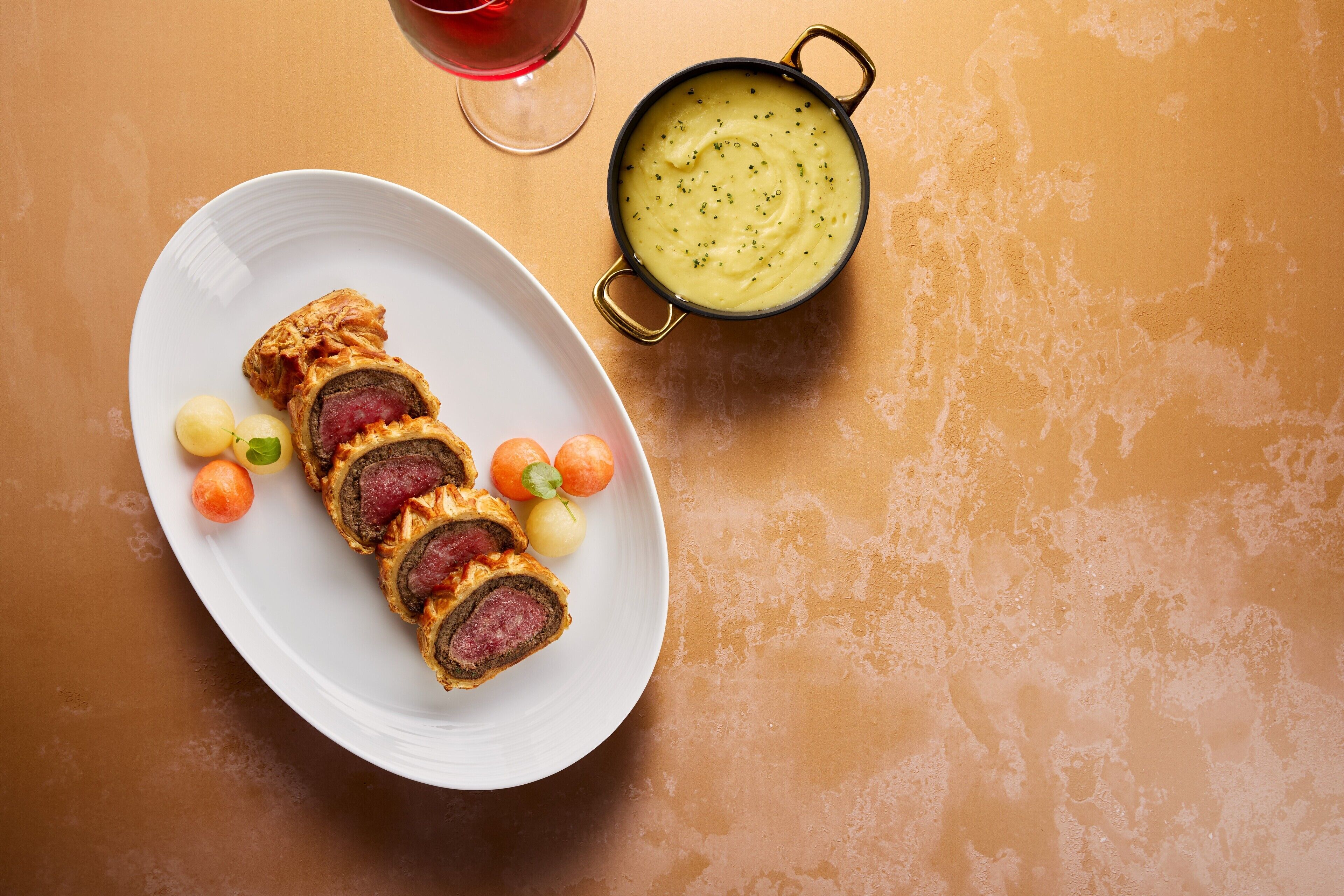 A plate with sliced meat rolls and small round fruits, a bowl of soup, and a glass of red wine on a textured orange surface.