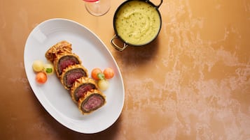 A plate with sliced meat rolls and small round fruits, a bowl of soup, and a glass of red wine on a textured orange surface.