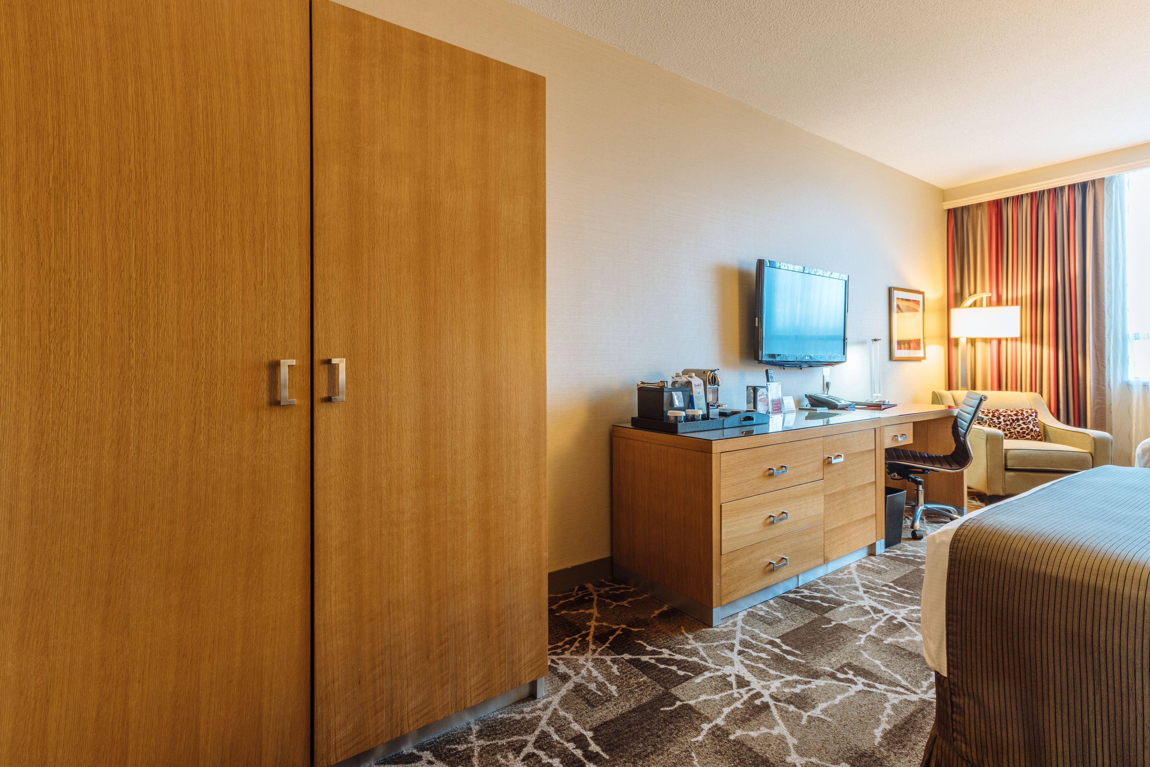A hotel room with a wooden wardrobe, a desk with a television, a chair, and a bed.