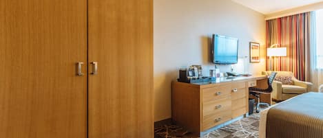 A hotel room with a wooden wardrobe, a desk with a television, a chair, and a bed.