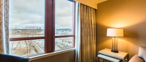A hotel room with a large window, a bedside table with a lamp, a chair, and a patterned carpet.