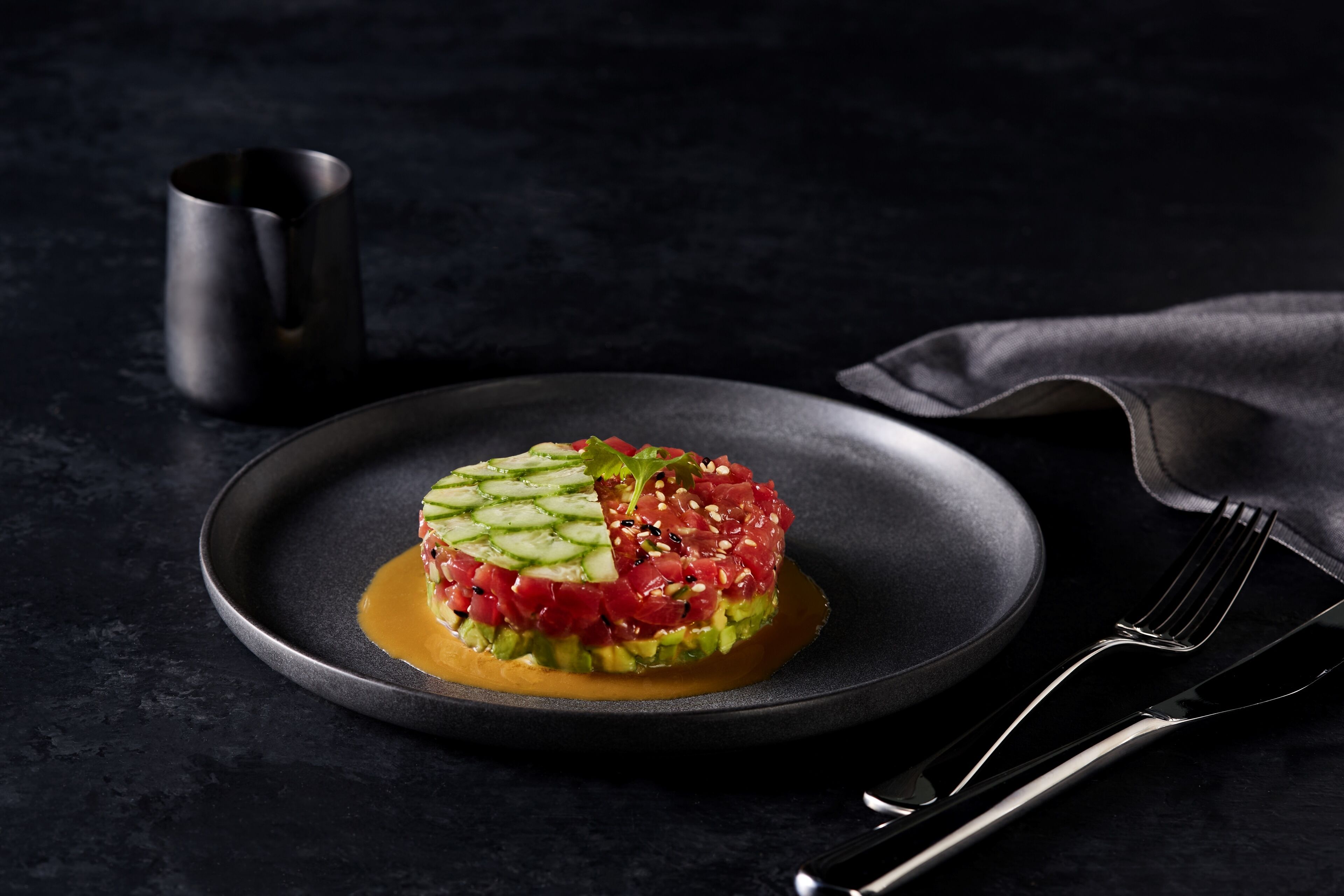 A dish of raw tuna tartare on a dark plate with a fork and a cup beside it.