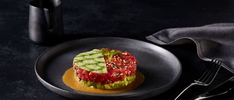 Un plato de tartare de atún crudo sobre un plato oscuro, con un tenedor y una taza a un lado.