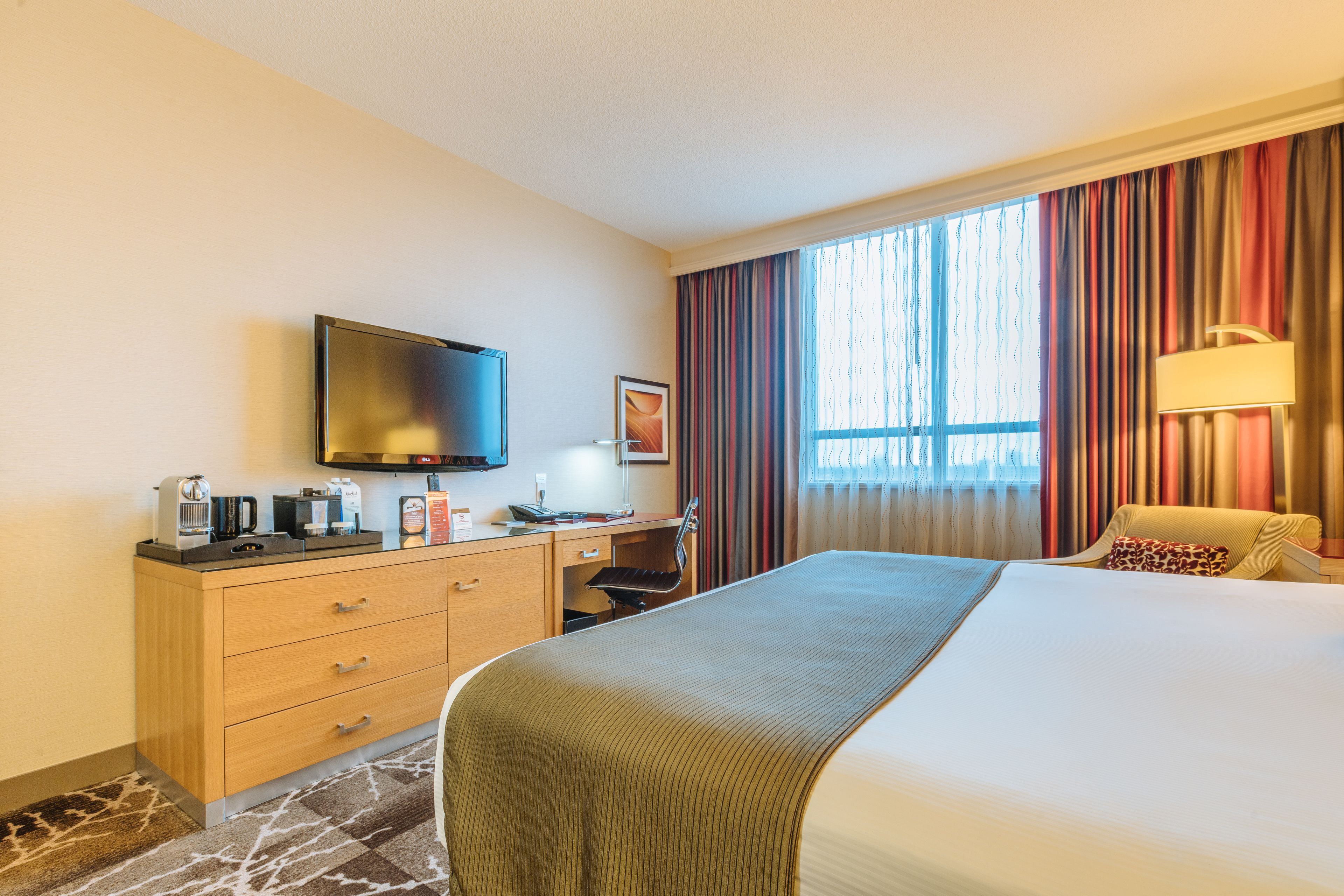 Quarto de hotel com uma cama, uma escrivaninha com televisão de tela plana, cafeteira e luminária.