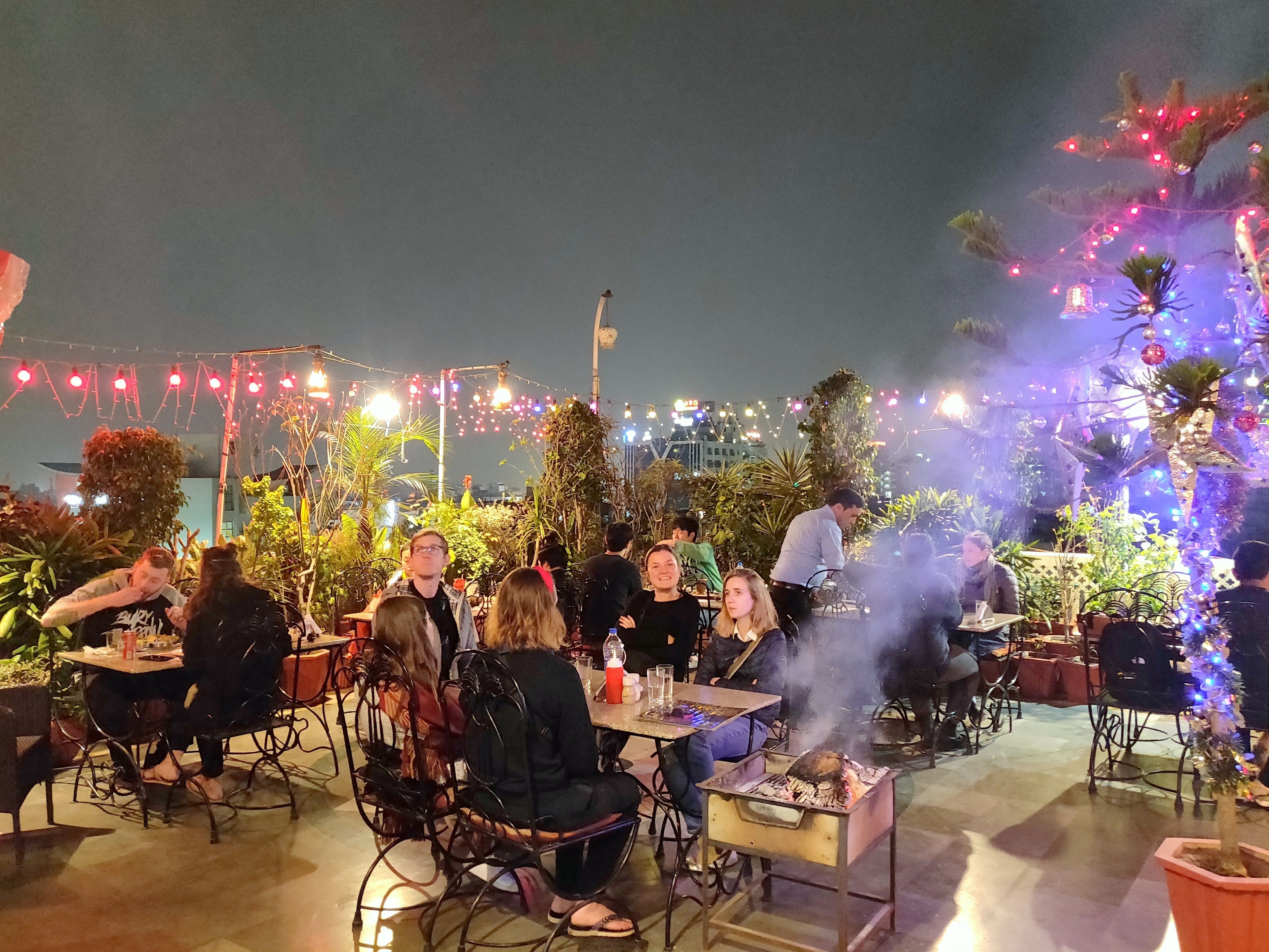 An outdoor dining area with tables and chairs, surrounded by potted plants and decorative lights.