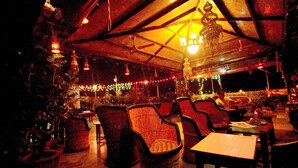 Een overdekte eetruimte met rieten stoelen en tafels, onder een groot, open dak met hangende lampen.