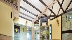 A restaurant with high ceilings, exposed beams, and a view of a waterfall through a glass wall. Tables are set with chairs, and there are water bottles on the tables. - Hotel Marble Arch (New Delhi)