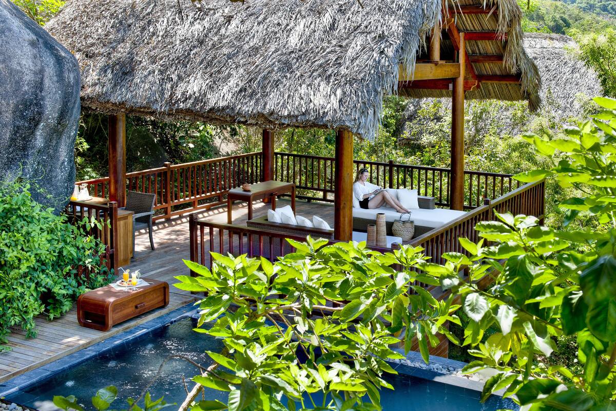 A thatched-roof outdoor lounge with wooden furniture, a small pool, and lush greenery.
