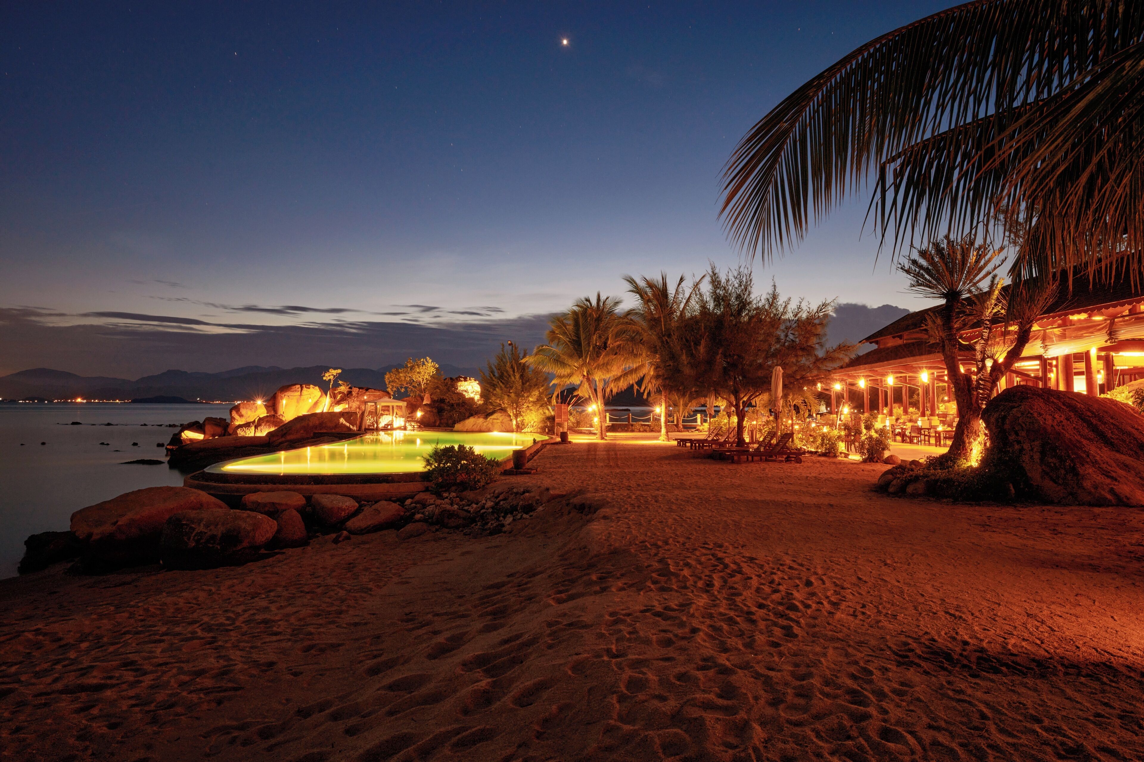 Un hotel frente a la playa con piscina, palmeras y vista al océano al atardecer.