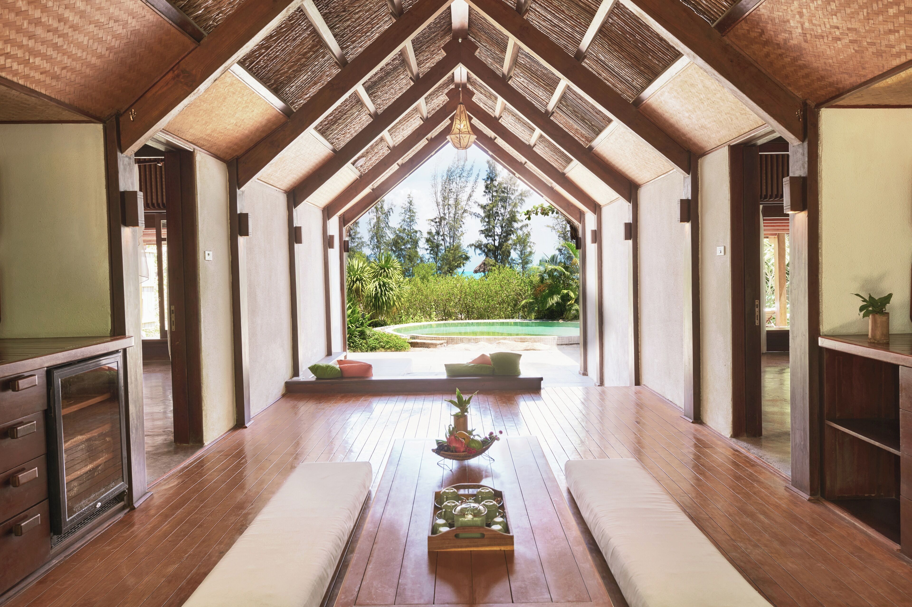 A spacious room with a wooden floor, a central seating area with cushions, and a view of a garden through a glass door.