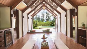 A spacious room with a wooden floor, a central seating area with cushions, and a view of a garden through a glass door.