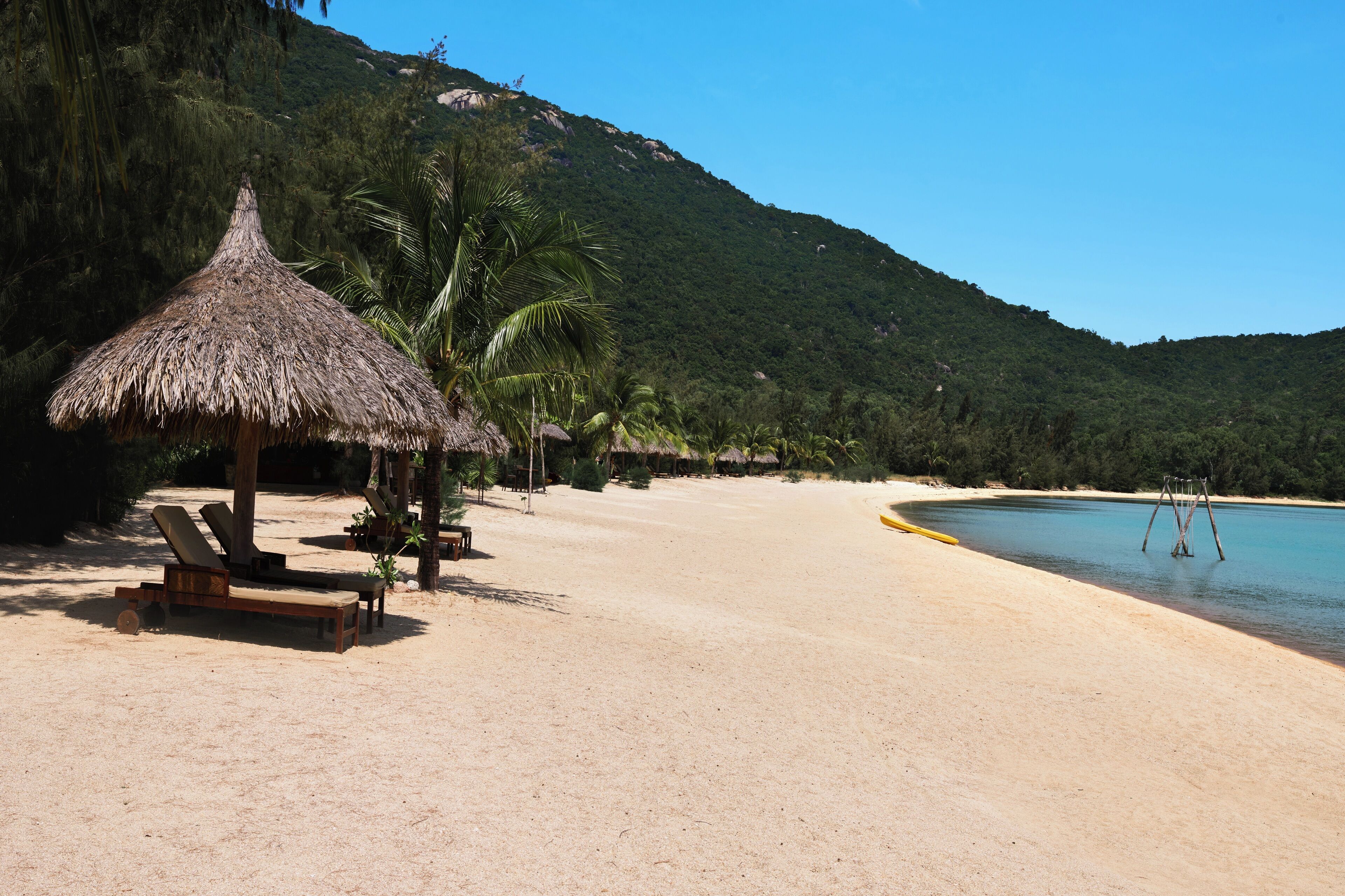 A beach with a thatched hut, lounge chairs, and a clear view of the ocean and a mountain in the background.