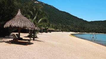 A beach with a thatched hut, lounge chairs, and a clear view of the ocean and a mountain in the background.