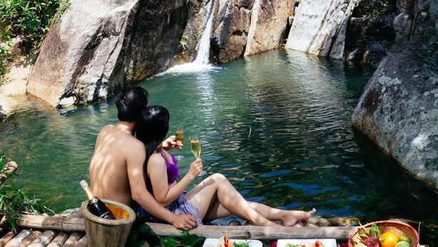 Una pareja sentada en una terraza de madera junto a una cascada, con comida y bebidas en la mesa.