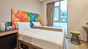 Ein Hotelzimmer mit zwei Betten, einem Schreibtisch mit Kaffeemaschine, einem Kühlschrank, einem farbenfrohen abstrakten Wandbild, einem Fenster mit Vorhängen und einem kleinen Hocker.