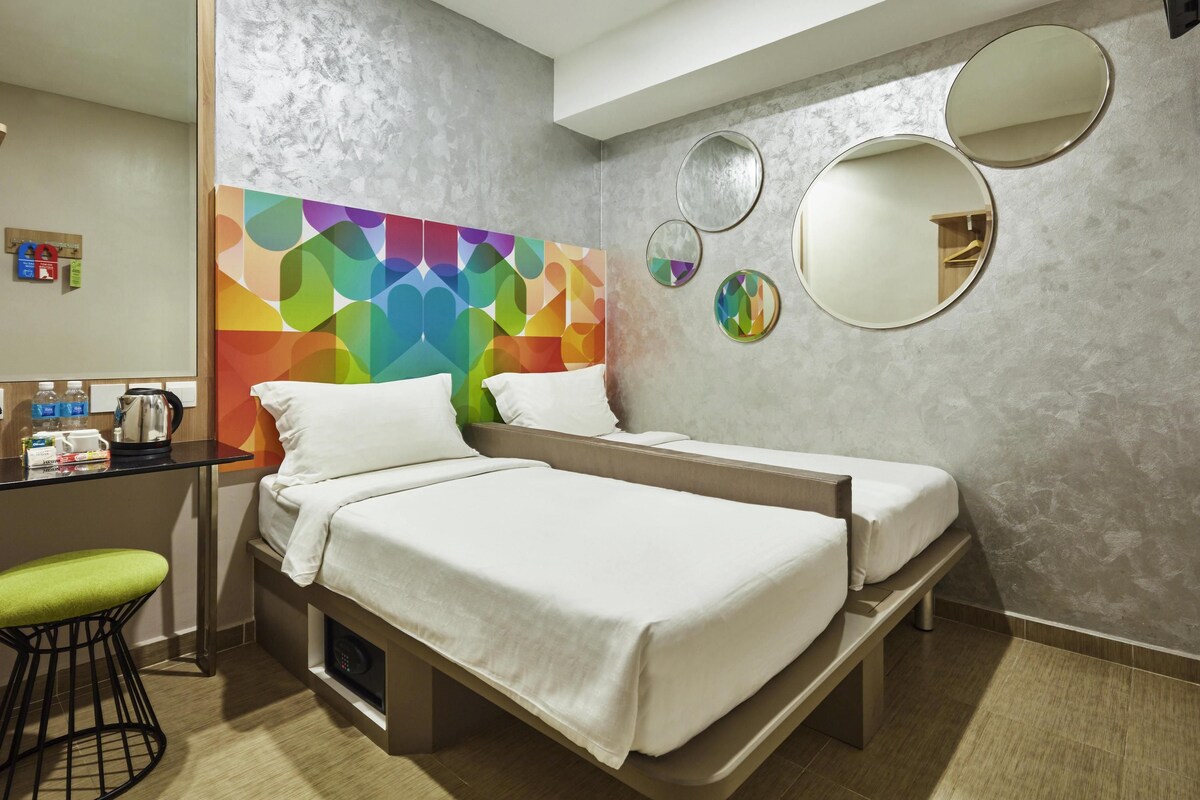 A hotel room with two beds, a desk with a kettle, a chair, and a colorful geometric wall design.