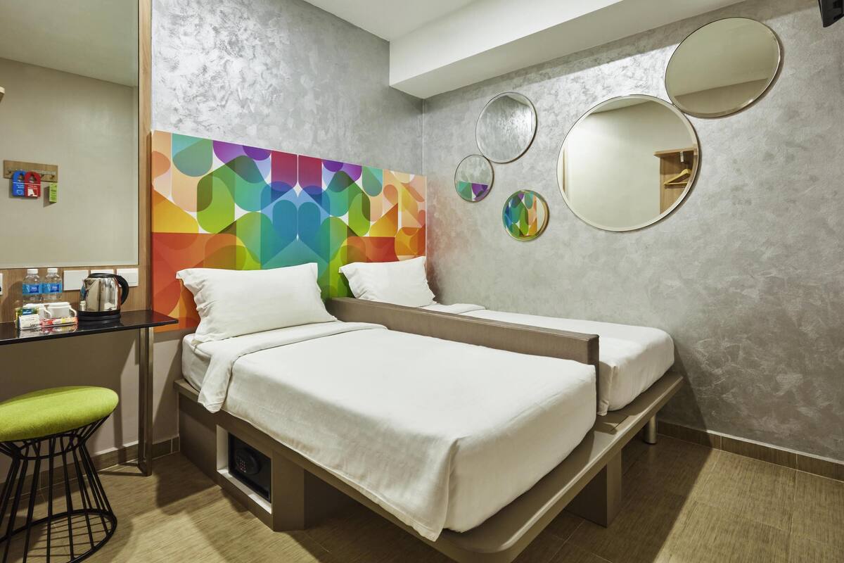 A hotel room with two beds, a desk with a kettle, a chair, and a colorful geometric wall design.