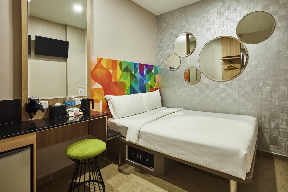 A hotel room with a bed, a desk with a chair, a mirror, and a colorful abstract wall art.