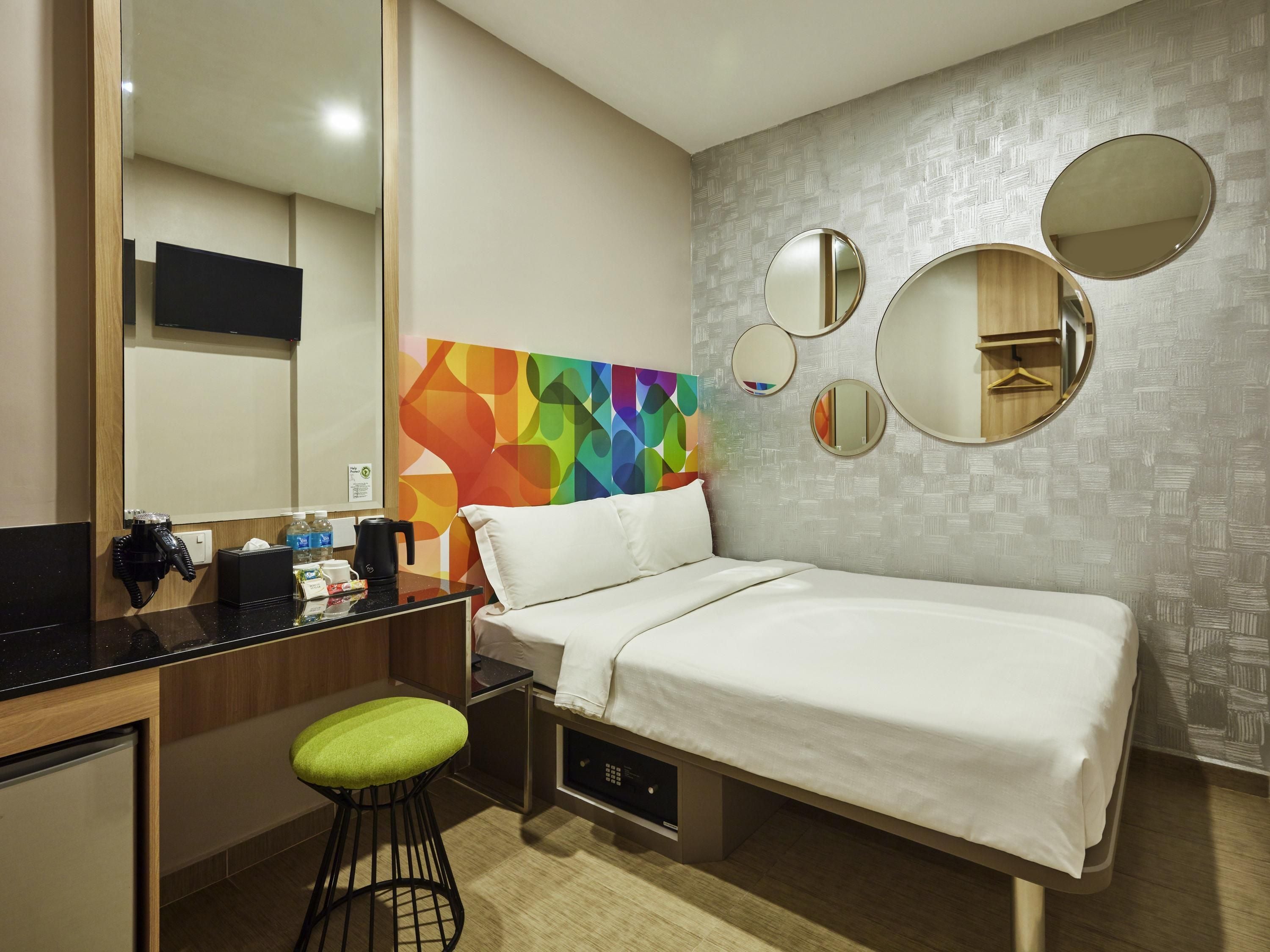 Une chambre d’hôtel comprenant un lit, un bureau avec une chaise, un miroir et une œuvre d’art murale abstraite aux couleurs vives.