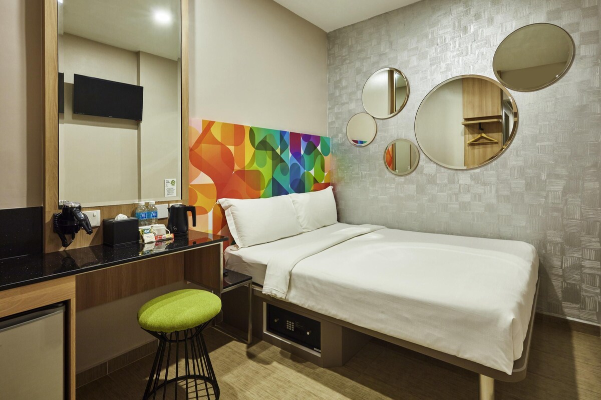 A hotel room with a bed, a desk with a chair, a mirror, and a colorful abstract wall art.