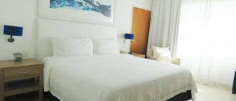 A hotel room with a large bed, a bedside table, a nightstand, a chair, a desk, a lamp, and a painting on the wall.