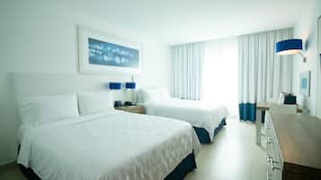 A hotel room with two beds, a desk, and a chair. There is a window with curtains and a painting on the wall.