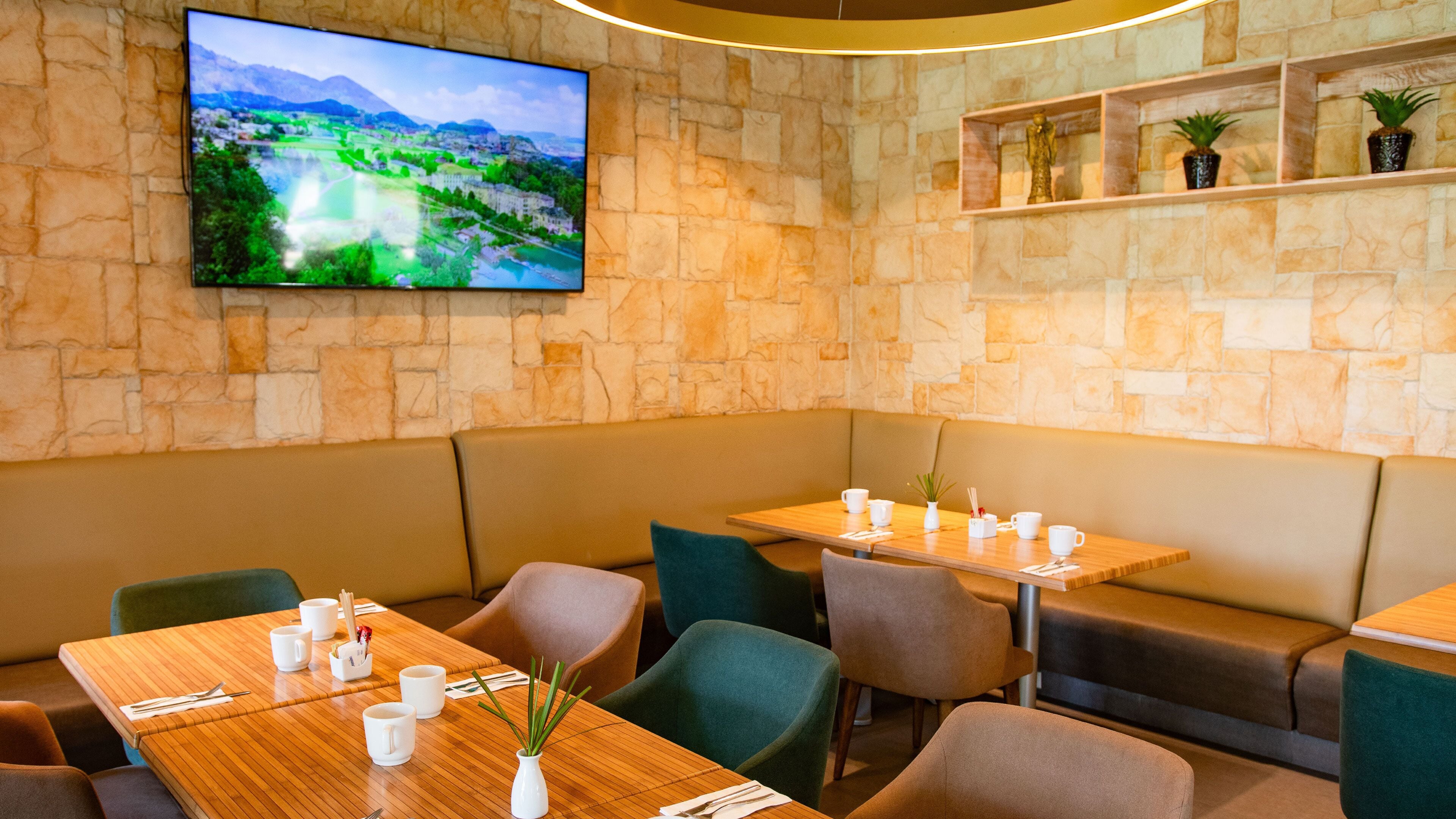 A restaurant with stone walls, green cushioned seating, wooden tables, and a flat-screen TV displaying a landscape.