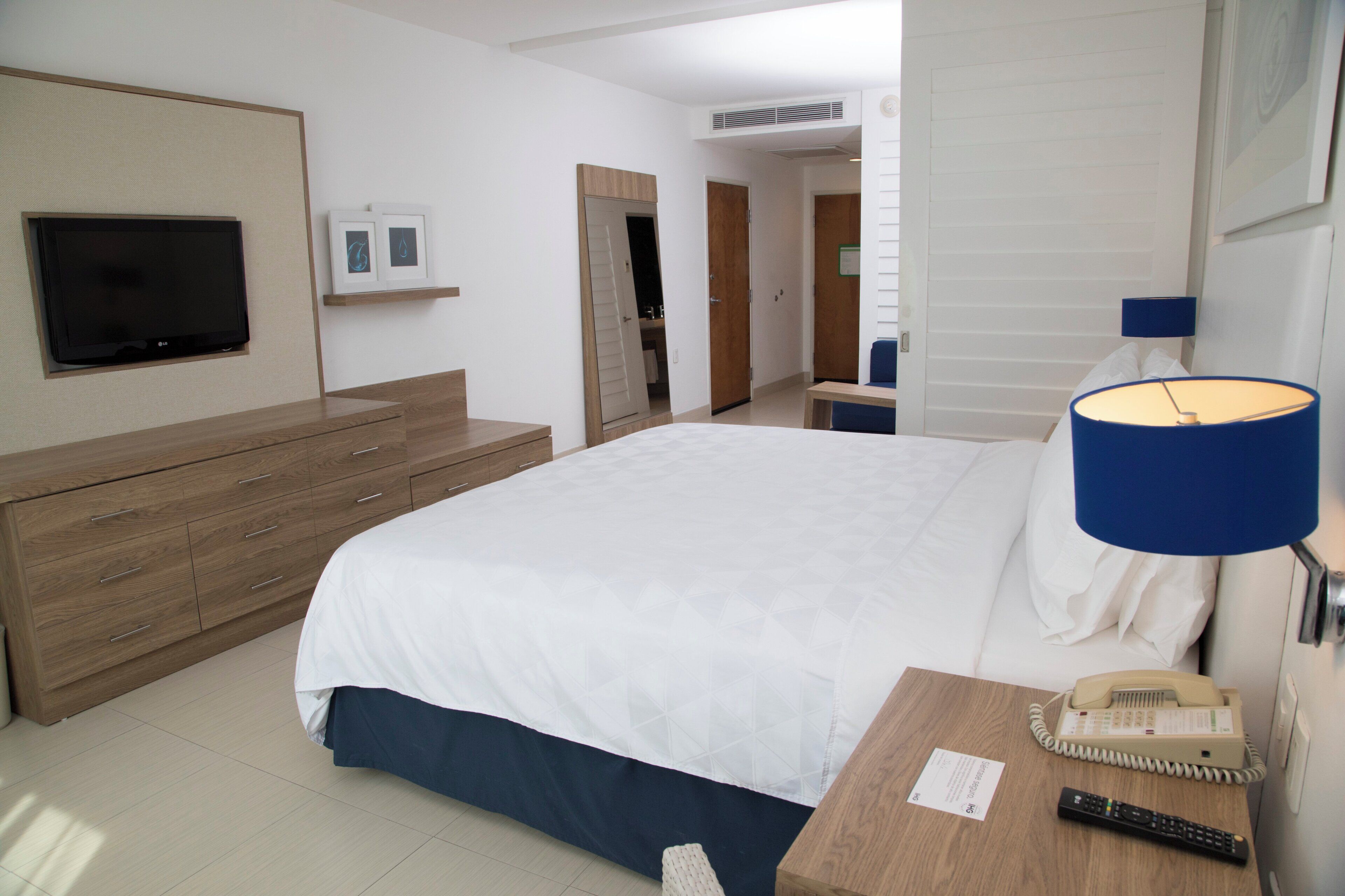 A hotel room with a bed, a TV mounted on the wall, a bedside table with a lamp, a phone, and a remote control.
