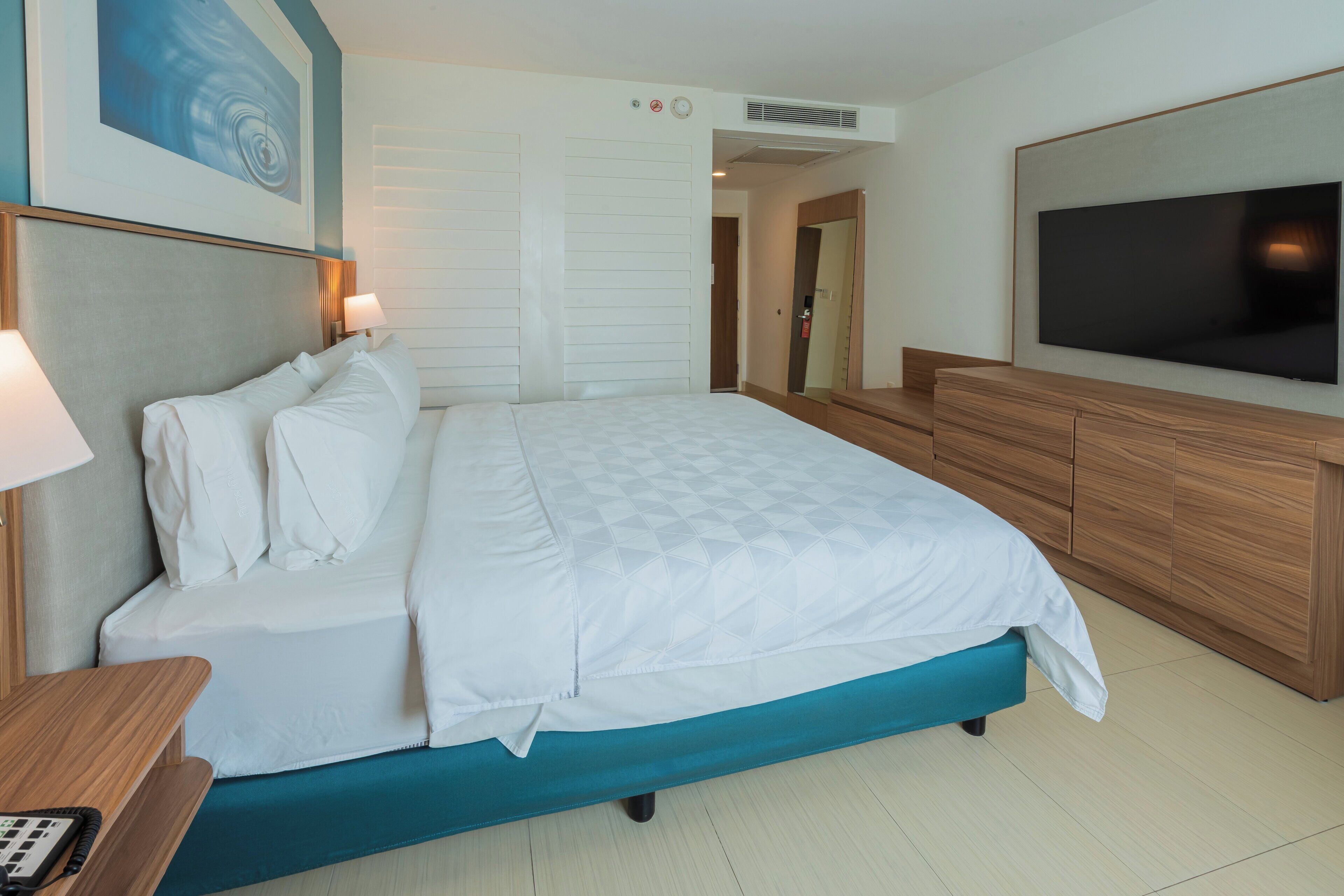 A hotel room with a bed, a TV, a desk, and a painting on the wall.