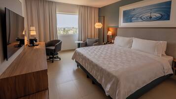 Habitación de hotel con una cama grande, un escritorio con televisor, una silla y vista a la ciudad por la ventana.