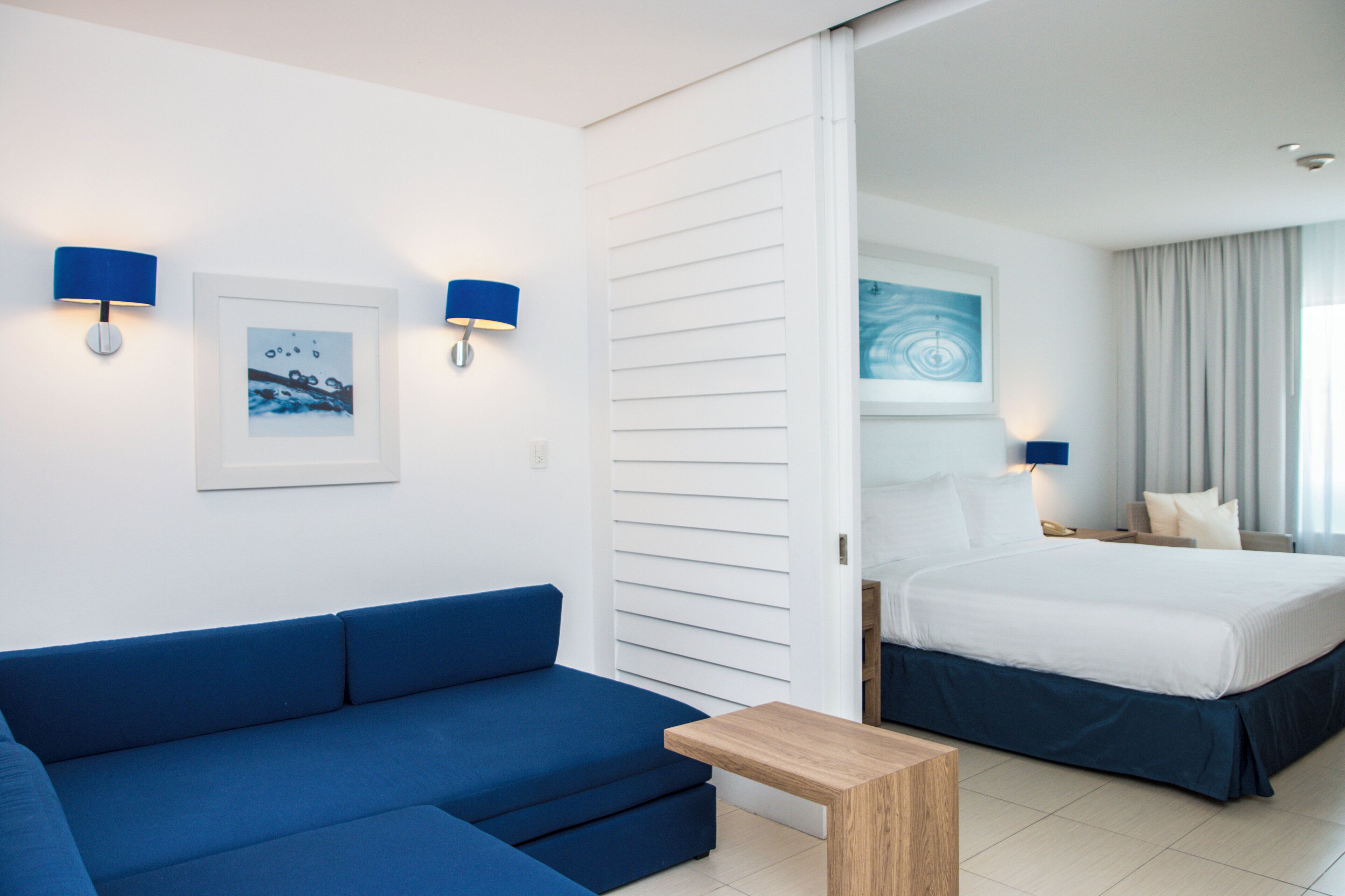 Een hotelkamer met een blauwe bank, een houten bijzettafeltje, een bed met wit bedgoed en twee wandlampen.