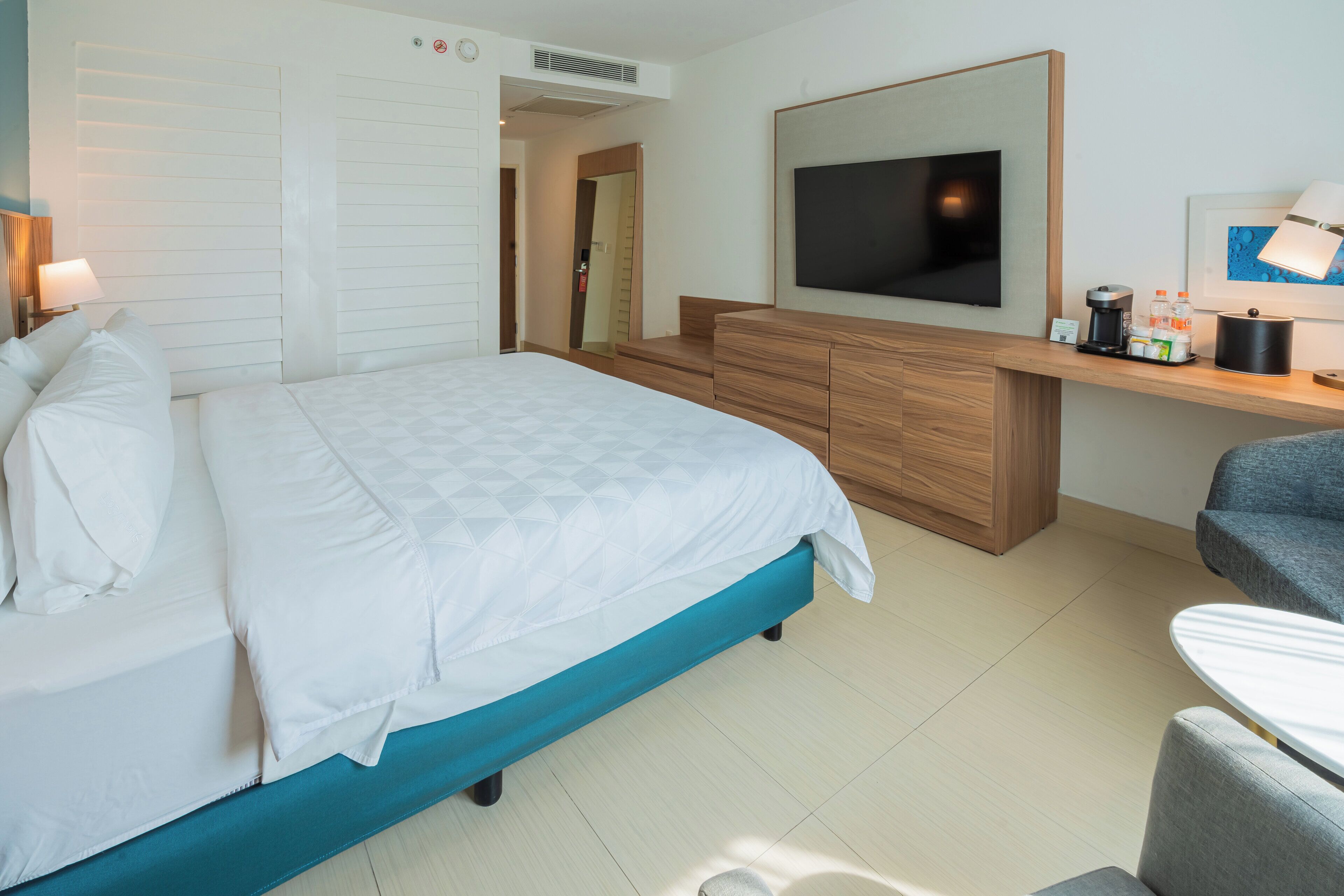 A modern hotel room with a large bed, a TV, a desk with a lamp, and a coffee machine.