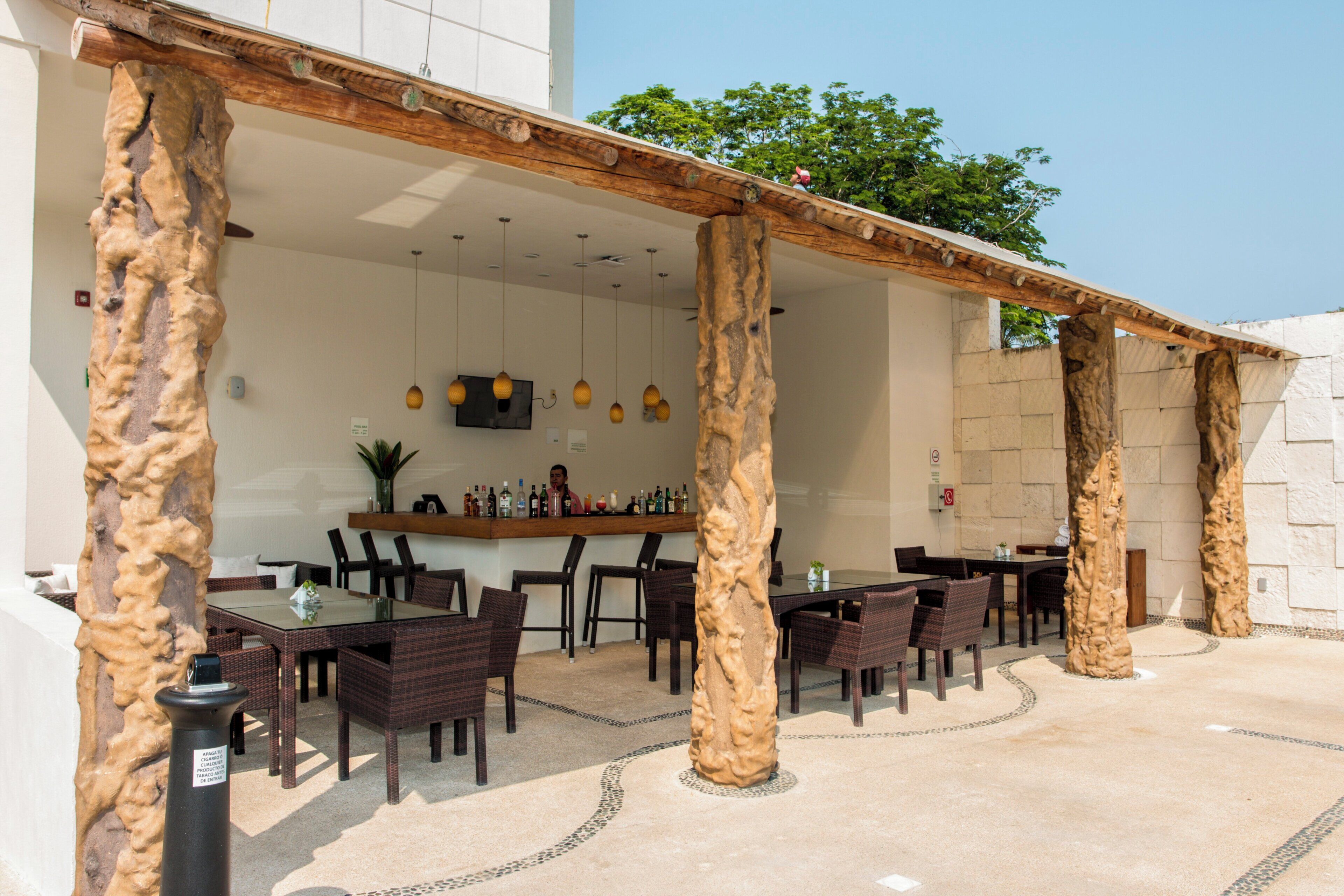 Un espacio al aire libre con mesas y sillas, una barra con botellas y una estructura de pilares de piedra.