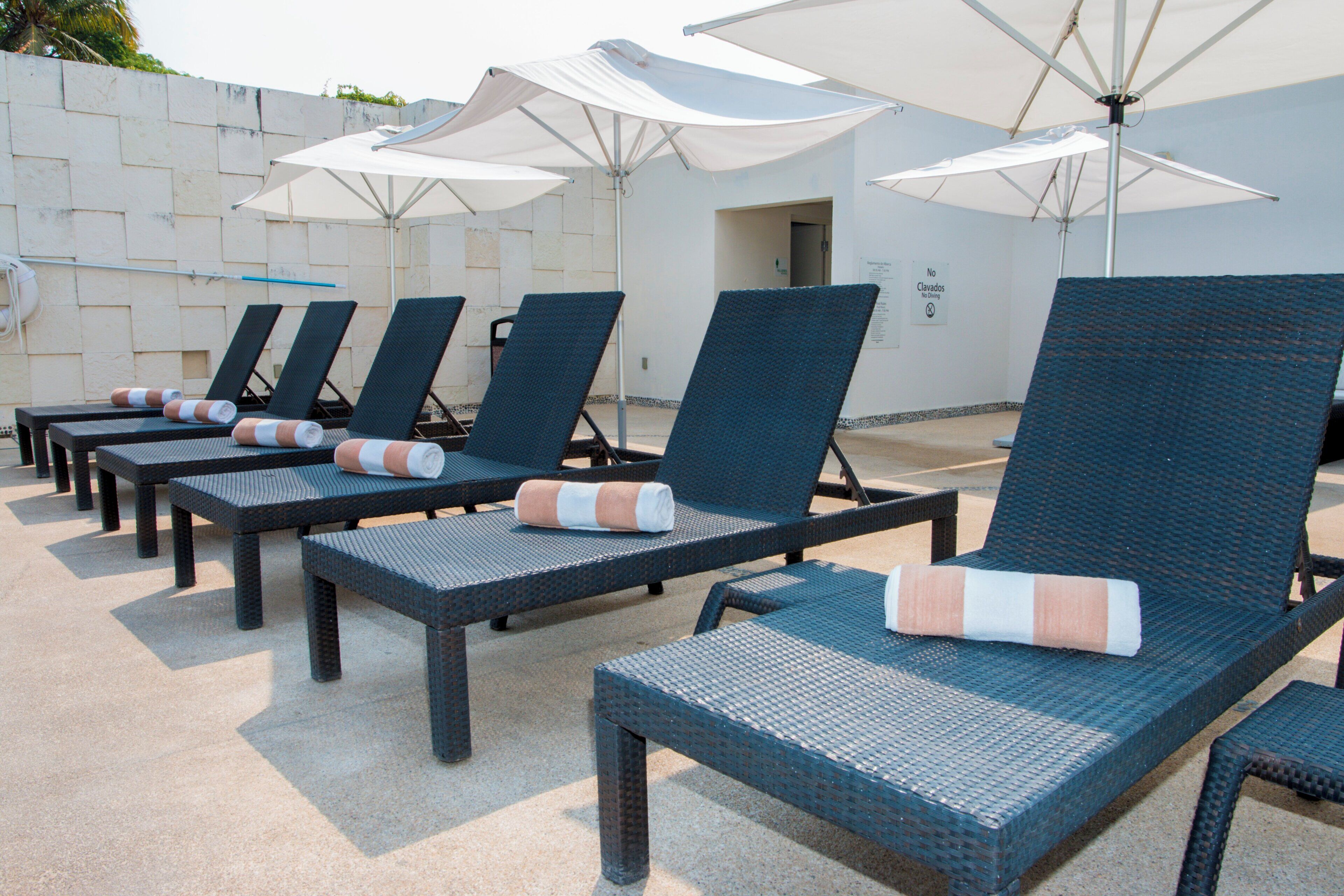 A row of lounge chairs with cushions, arranged under umbrellas, against a wall with a sign.