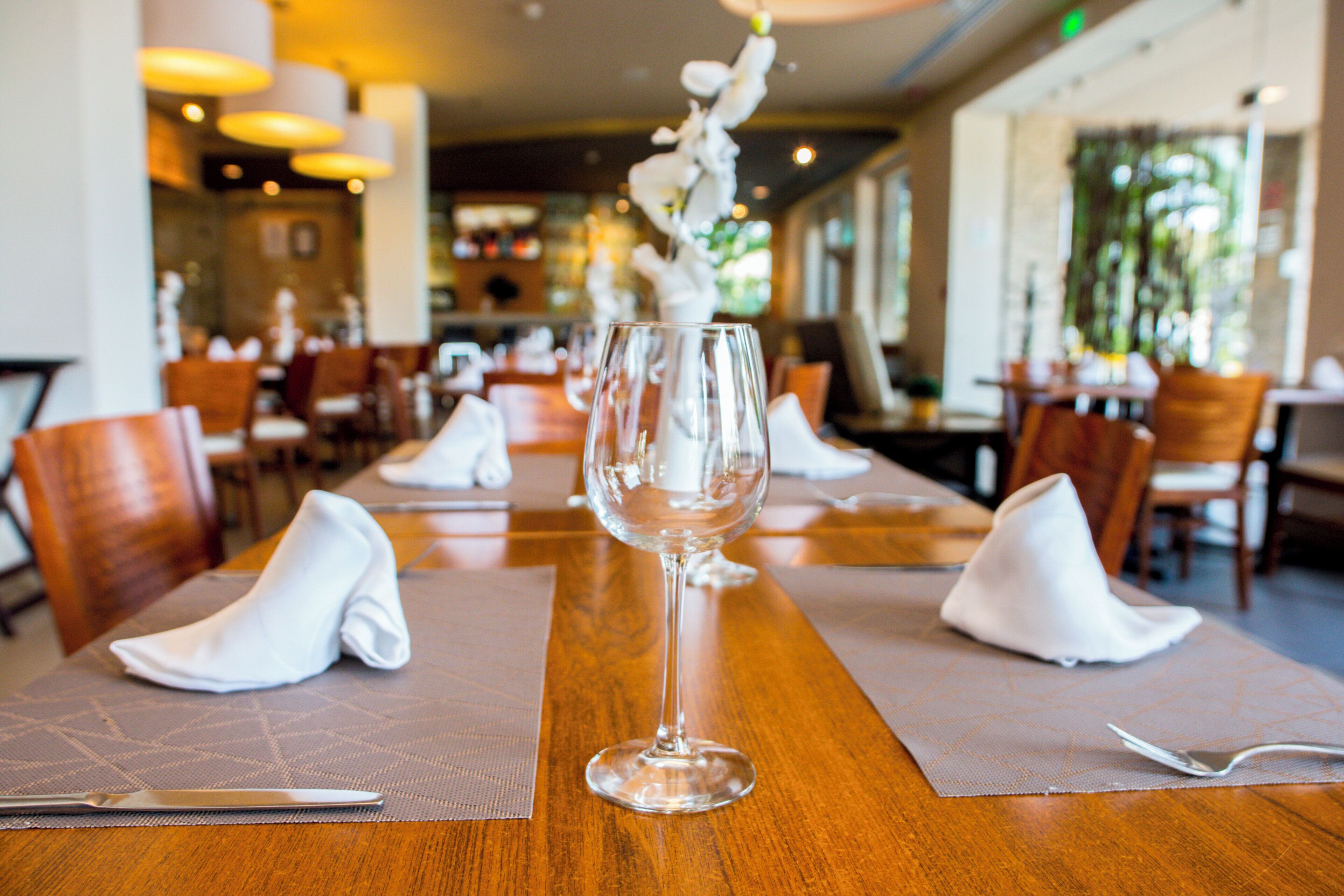 A restaurant setting with a long wooden table, white napkins, and a clear glass wine glass.