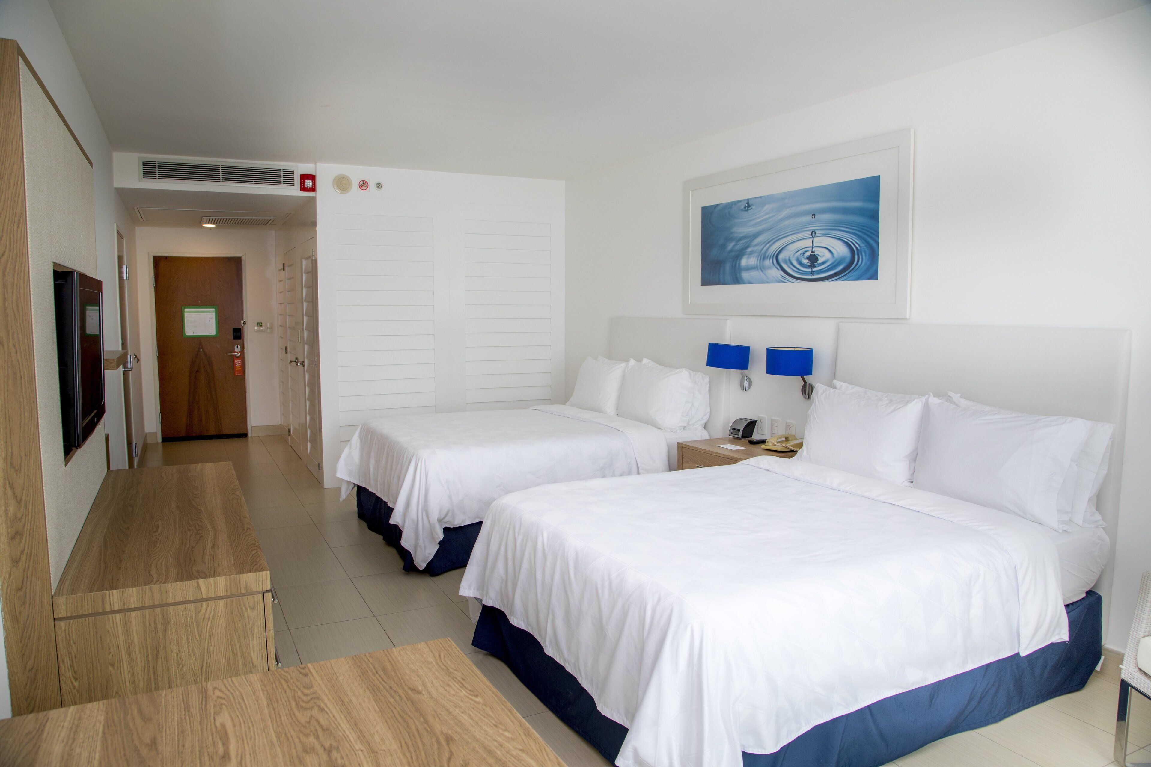 A hotel room with two beds, a desk, a chair, a TV, and a painting on the wall.