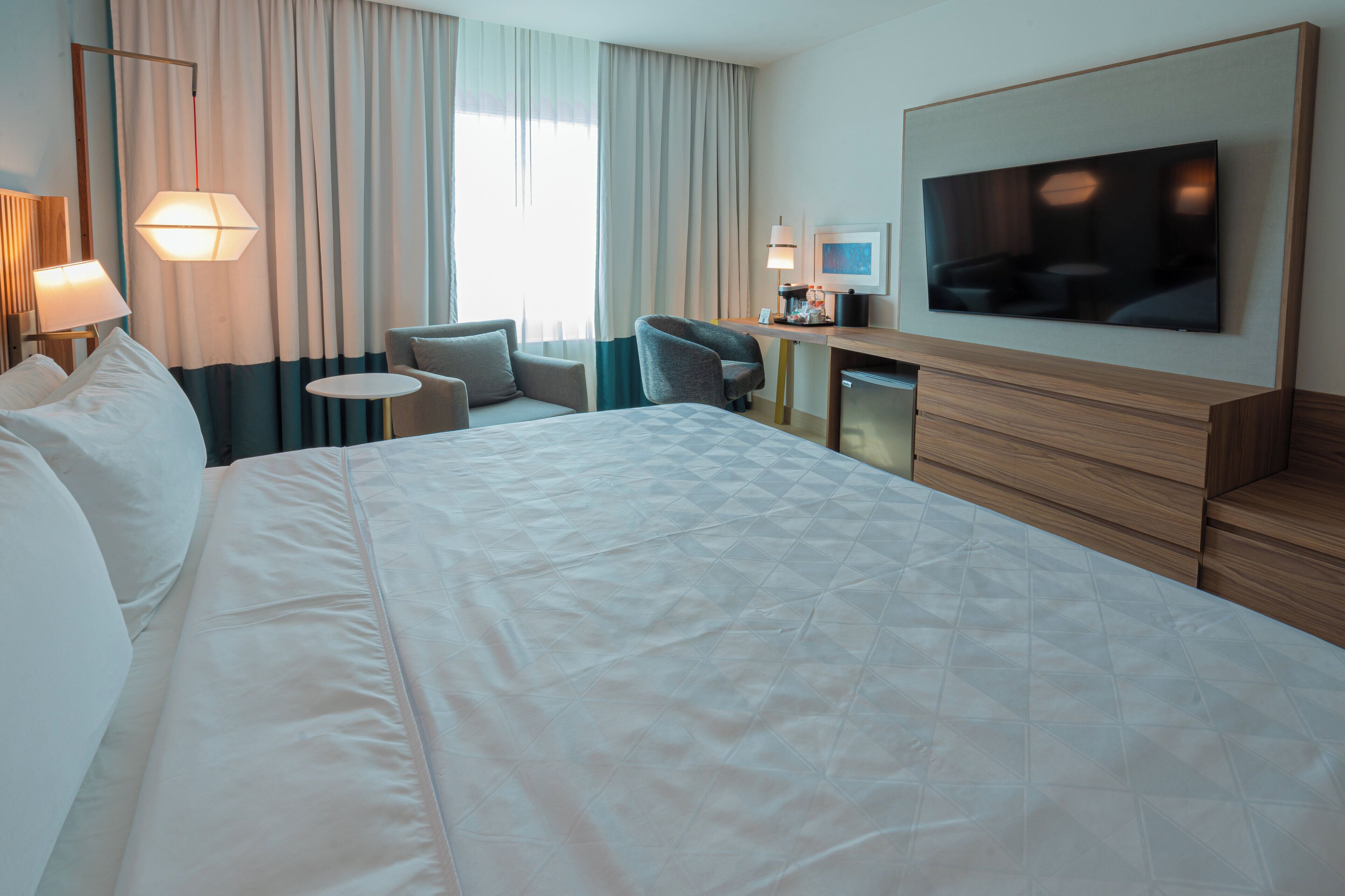 Habitación de hotel con una cama grande, un escritorio con televisor de pantalla plana, una silla, una mesita pequeña y una lámpara.