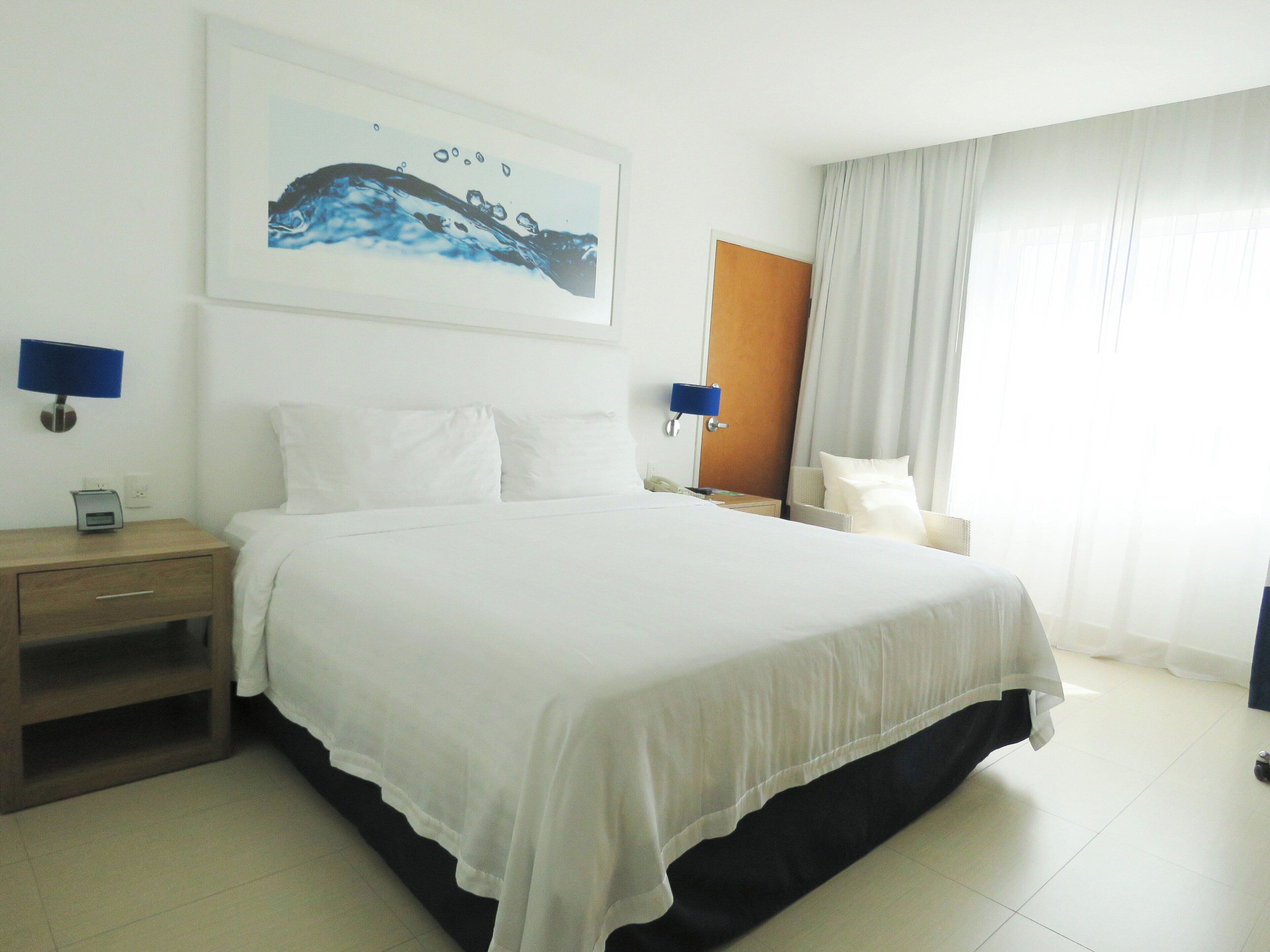 A hotel room with a large bed, a nightstand, a desk, a chair, and a painting on the wall.