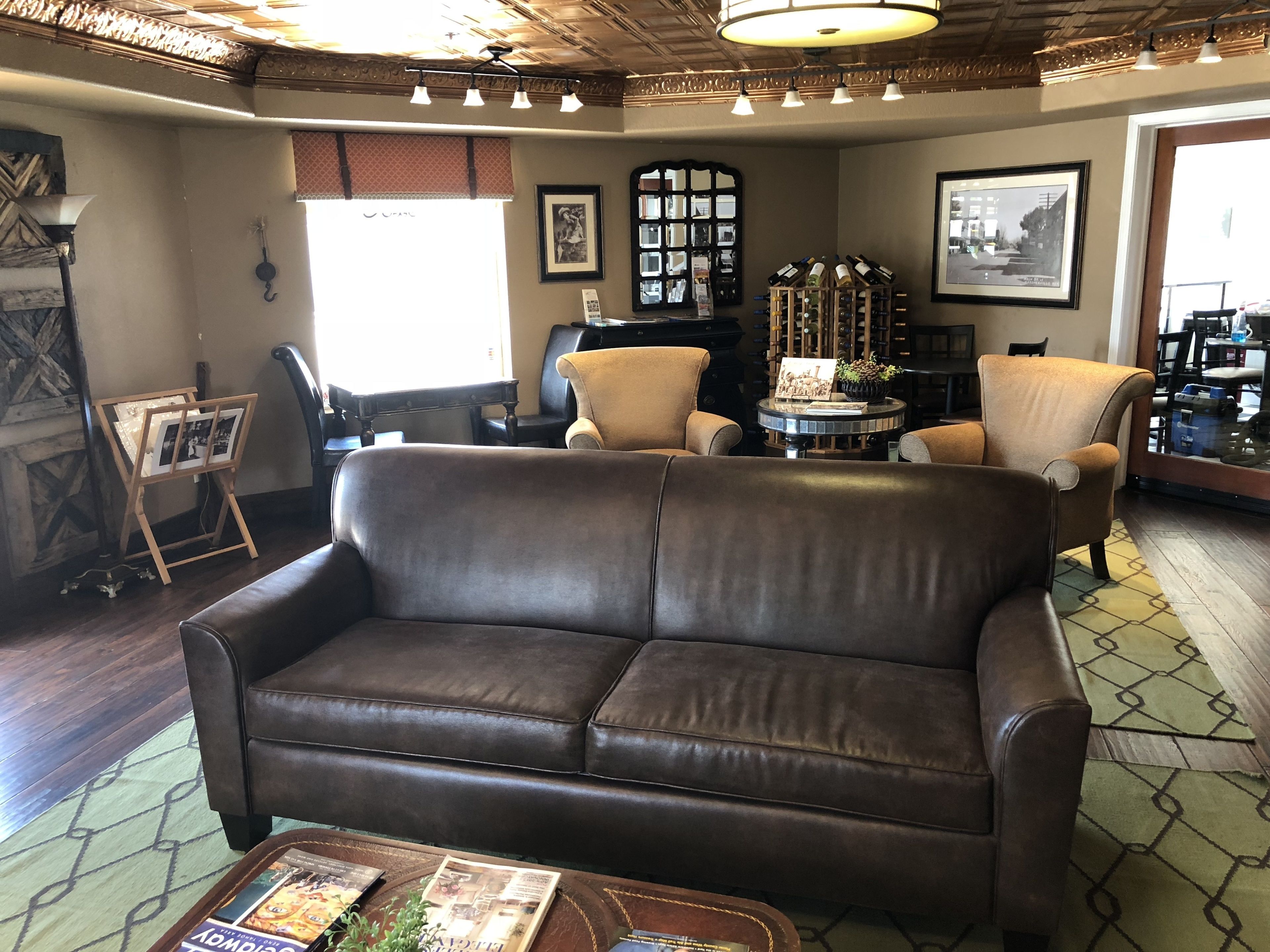 A cozy living room with a brown leather sofa, a small round table, and a coffee table with magazines. There are two armchairs, a fireplace, and a wine rack with bottles.