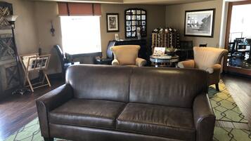 A cozy living room with a brown leather sofa, a small round table, and a coffee table with magazines. There are two armchairs, a fireplace, and a wine rack with bottles.