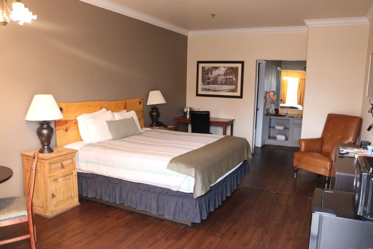 Une chambre d’hôtel avec un grand lit, deux lampes de chevet, une table de chevet en bois, un bureau avec une chaise, un fauteuil en cuir marron et un tableau encadré au mur.