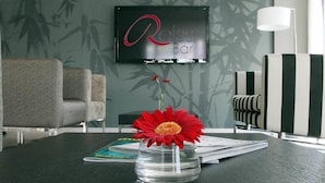 A modern hotel room with a flat-screen TV displaying "Reiss Bar" on the wall, a clear vase with a red flower on the table, and striped armchairs. - Golden Tulip Kassel Hotel Reiss (Kassel)