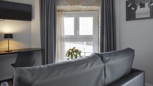 A modern living room with a grey sofa, a flat-screen TV mounted on the wall, a desk with a lamp, and a window with curtains. - Hotel Arrizul Beach (San Sebastián)