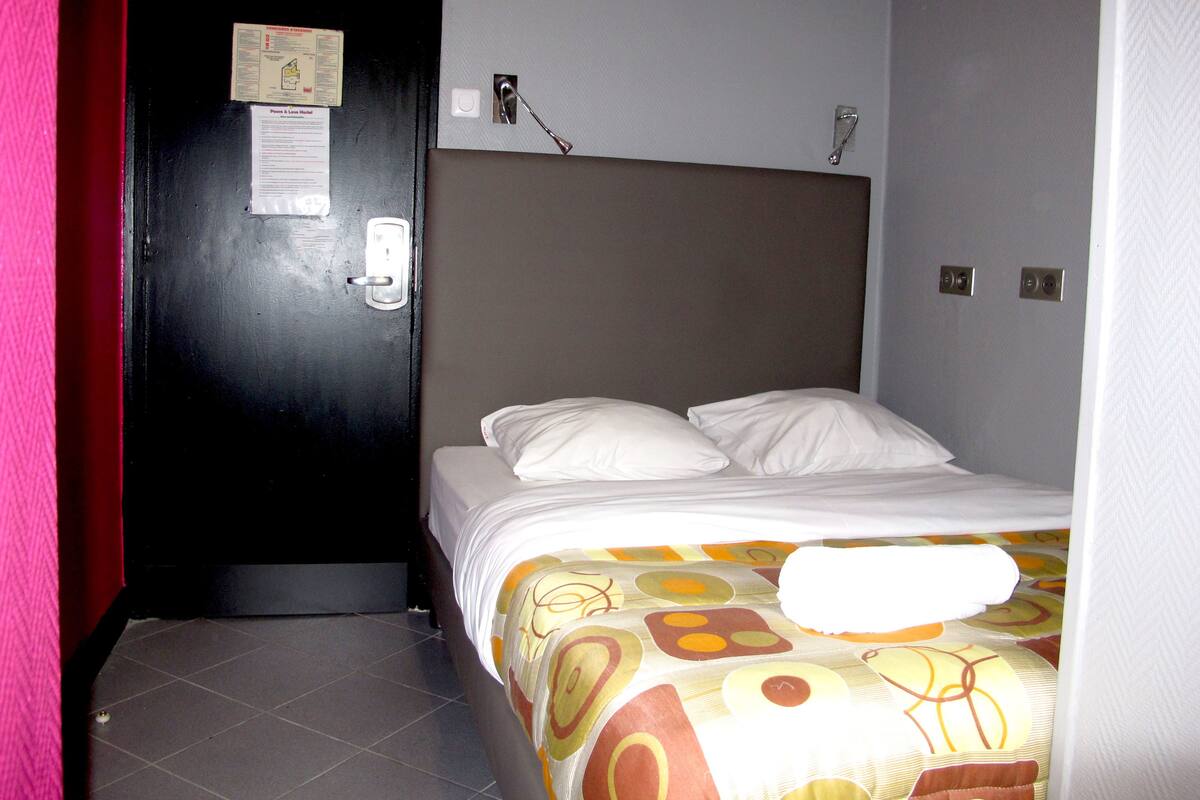 A hotel room with a bed, a door, and a wall-mounted light fixture.