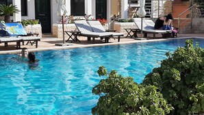 Una piscina con sillones de playa y sombrillas, rodeada por un edificio con ventanas de arco y fachada de piedra.
