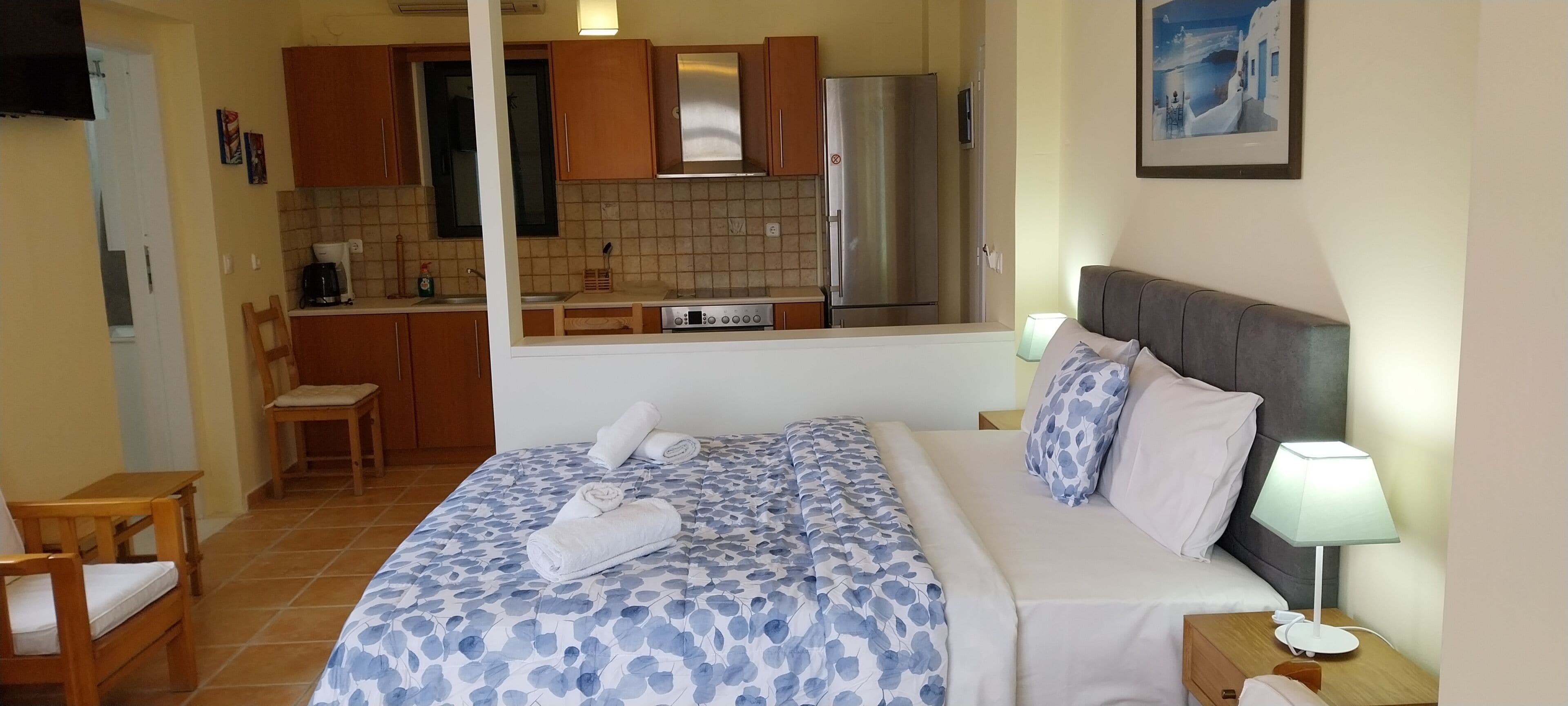 A bedroom with a bed, bedside table, lamp, and a view of a kitchen with cabinets and a refrigerator.